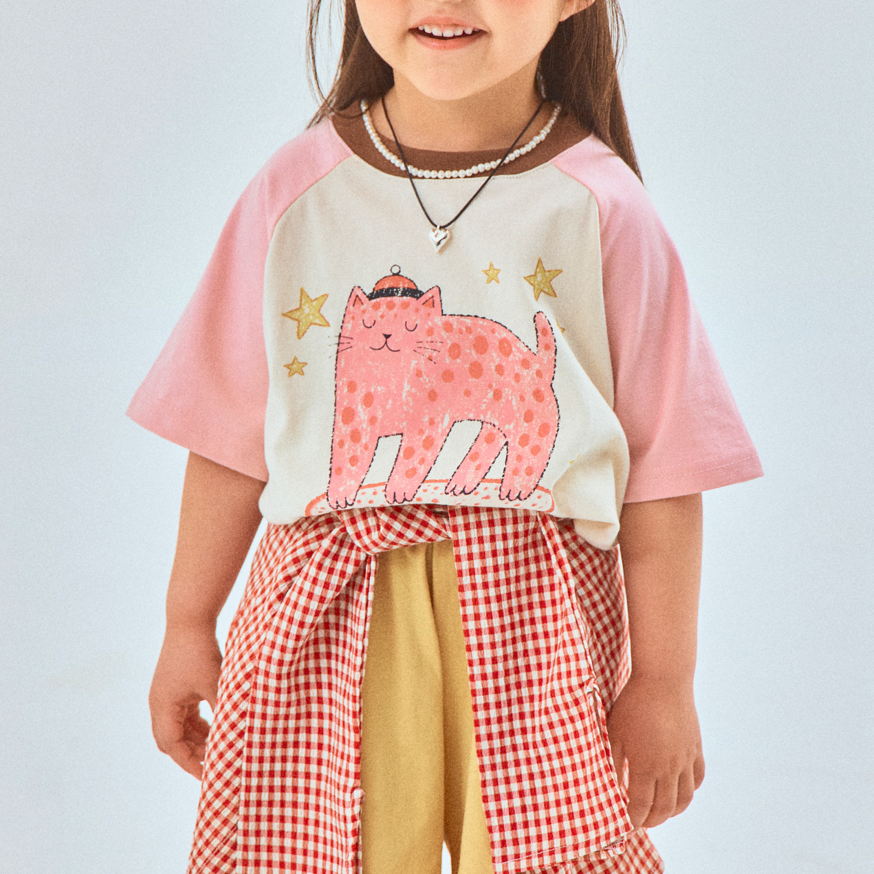 Child wearing a pink and white shirt with a cat design, yellow shorts, and a black cap on a light blue background.