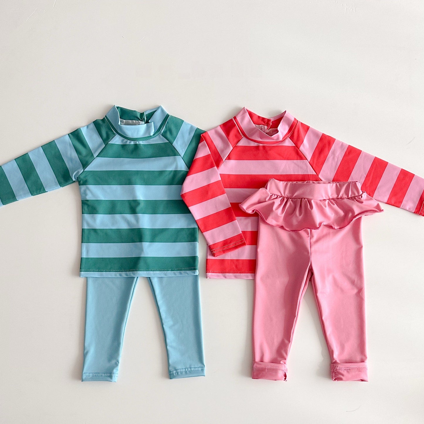 Two children's outfits, one green and blue striped shirt with matching pants, and one pink and red striped shirt with pink pants, on a light gray background.