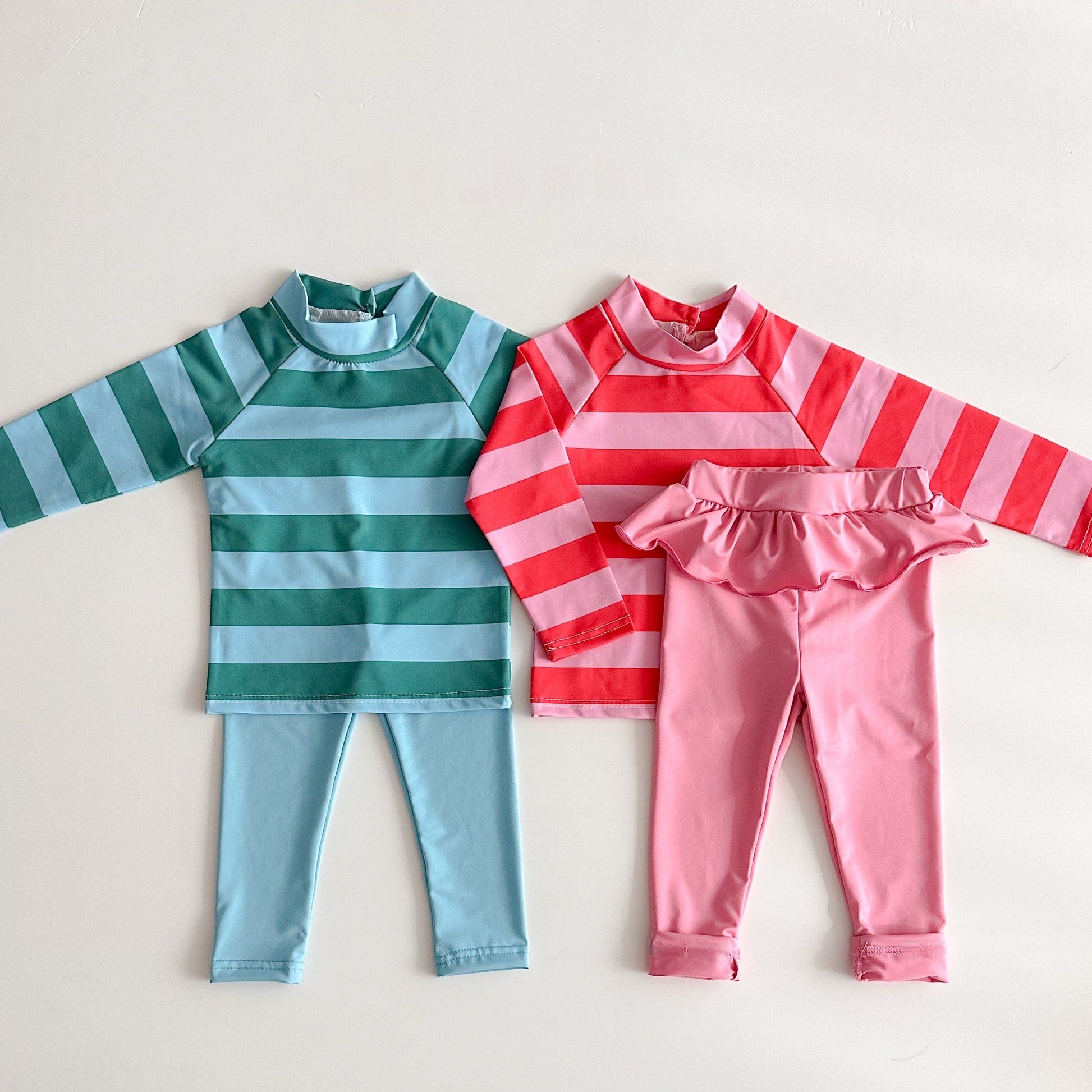 Two children's outfits, one green and blue striped shirt with matching pants, and one pink and red striped shirt with pink pants, on a light gray background.
